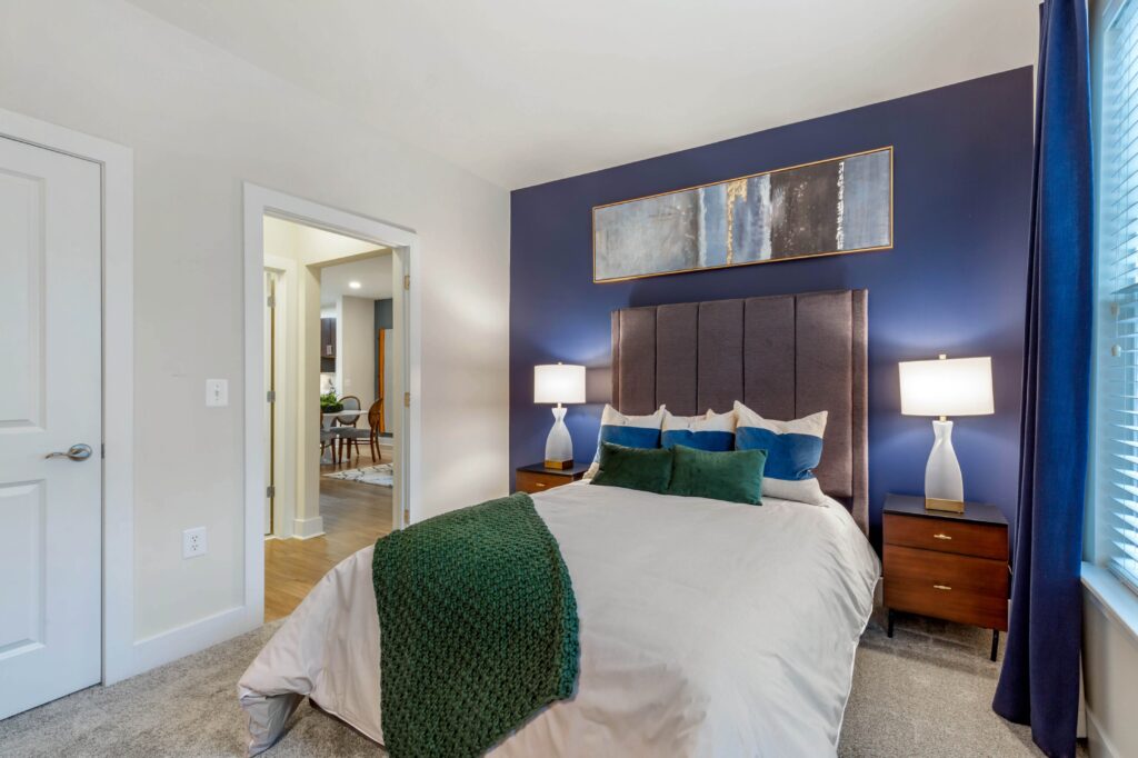 Bedroom with a dark blue accent wall, and carpet. Room for a queen size bed and two side tables.