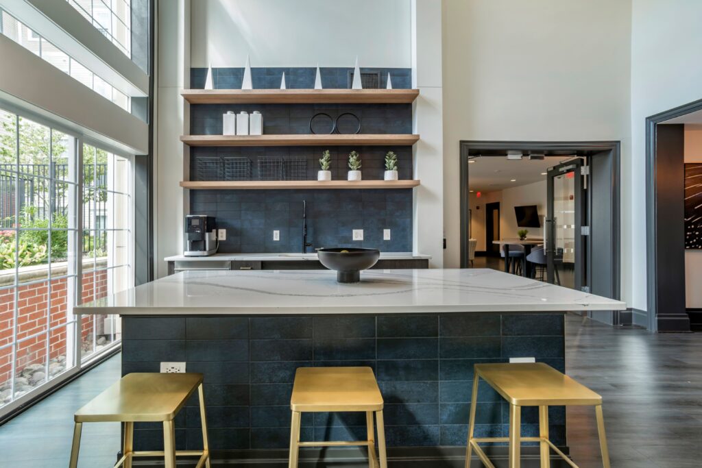 A small kichenette area in the clubhouse with bar seating for 3, a sink, and a coffee machine. There is an appliance under the counter but its unclear what it is.