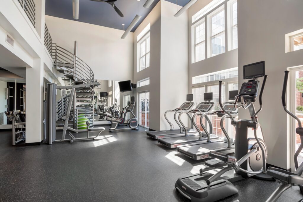 A two story fitness center with ample natural light and many machines including a cycle, bench, 3 treadmills and a stair machine with the implication that there is much more just out of view. TVs, screens on the machines, and a fan.