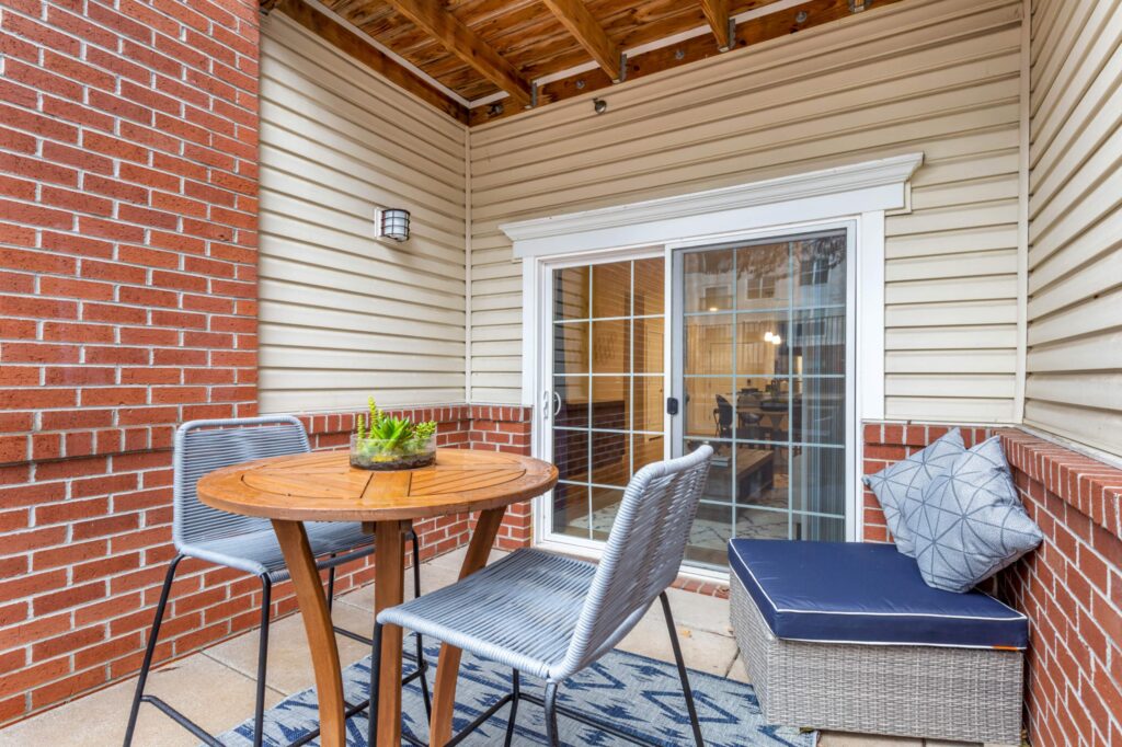 A balcony with big glass window doors, space for a table with 2 seats and a plush outdoor seat in the back. Siding and brick.